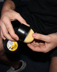 Person holding a black container with a yellow substance inside, against a dark background.