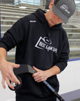 Person wearing a black hoodie with 'Best Dam Tup' logo and a gray cap, holding a hockey stick on an ice rink.